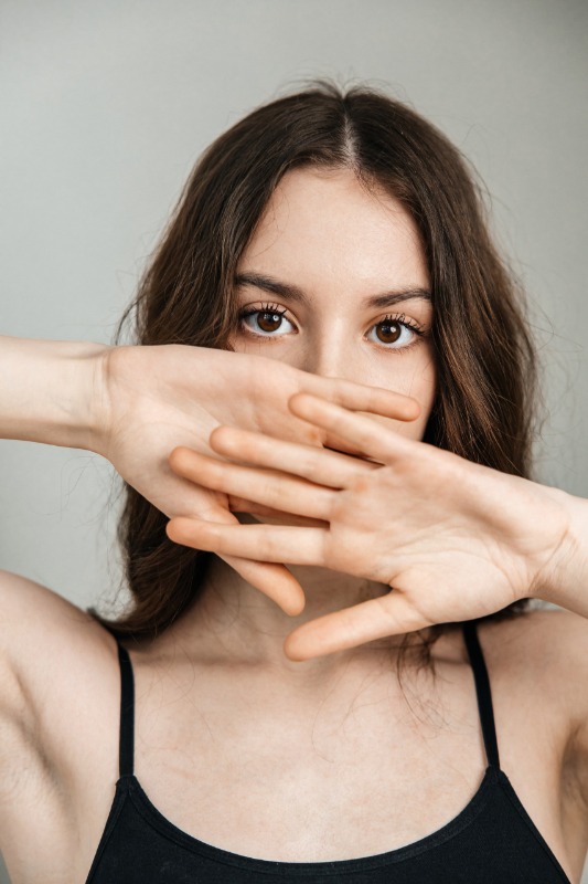 Девушка с темными волосами закрывает нижнюю часть лица скрещенными пальцами рук, смотрит в камеру