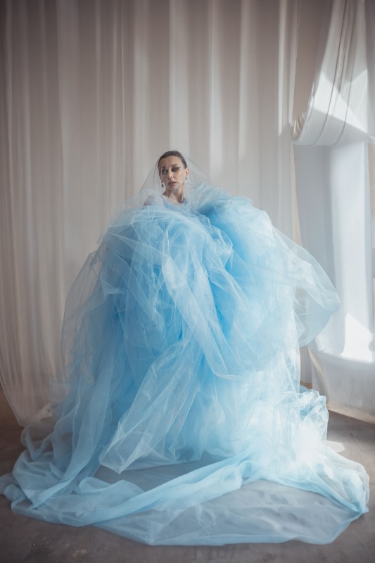 Женщина в пышном голубом платье из тюля позирует у окна с белыми шторами в светлом интерьере