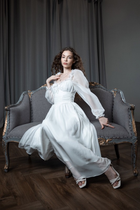 Женщина в длинном белом платье сидит на старинном сером диване с золотыми деталями на фоне серых штор