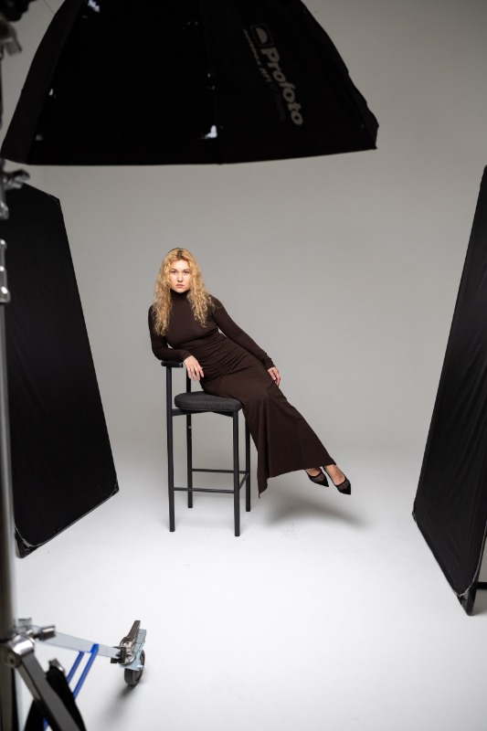 Женщина в коричневом платье сидит на высоком стуле в фотостудии с профессиональным освещением на белом фоне